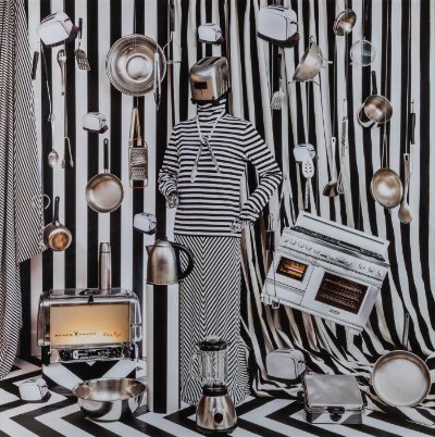 Figure in black and white striped clothing with a toaster head. There are black and white stripes behind them, and black and white chevron on the floor. They are surrounded by kitchen appliances.
